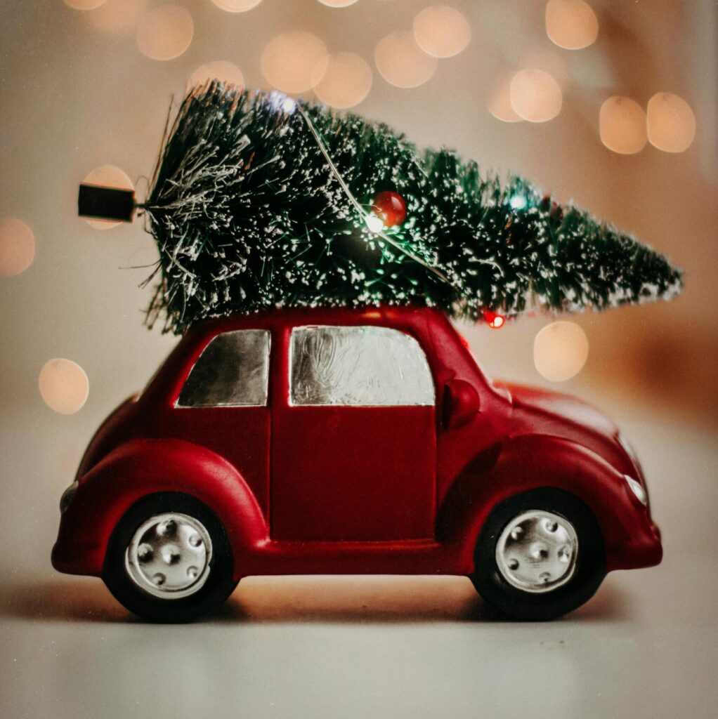 A red toy car carries a Christmas tree, set against a warm bokeh background, celebrating the festive season.